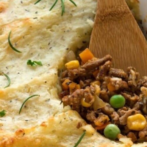 Shepherd's pie in a dish with a wooden spoon showcases a crowd-pleasing filling of ground meat, peas, corn, and carrots beneath mashed potatoes. Perfect for St. Patrick's Day celebrations, it's garnished with herbs to enhance its festive flair.