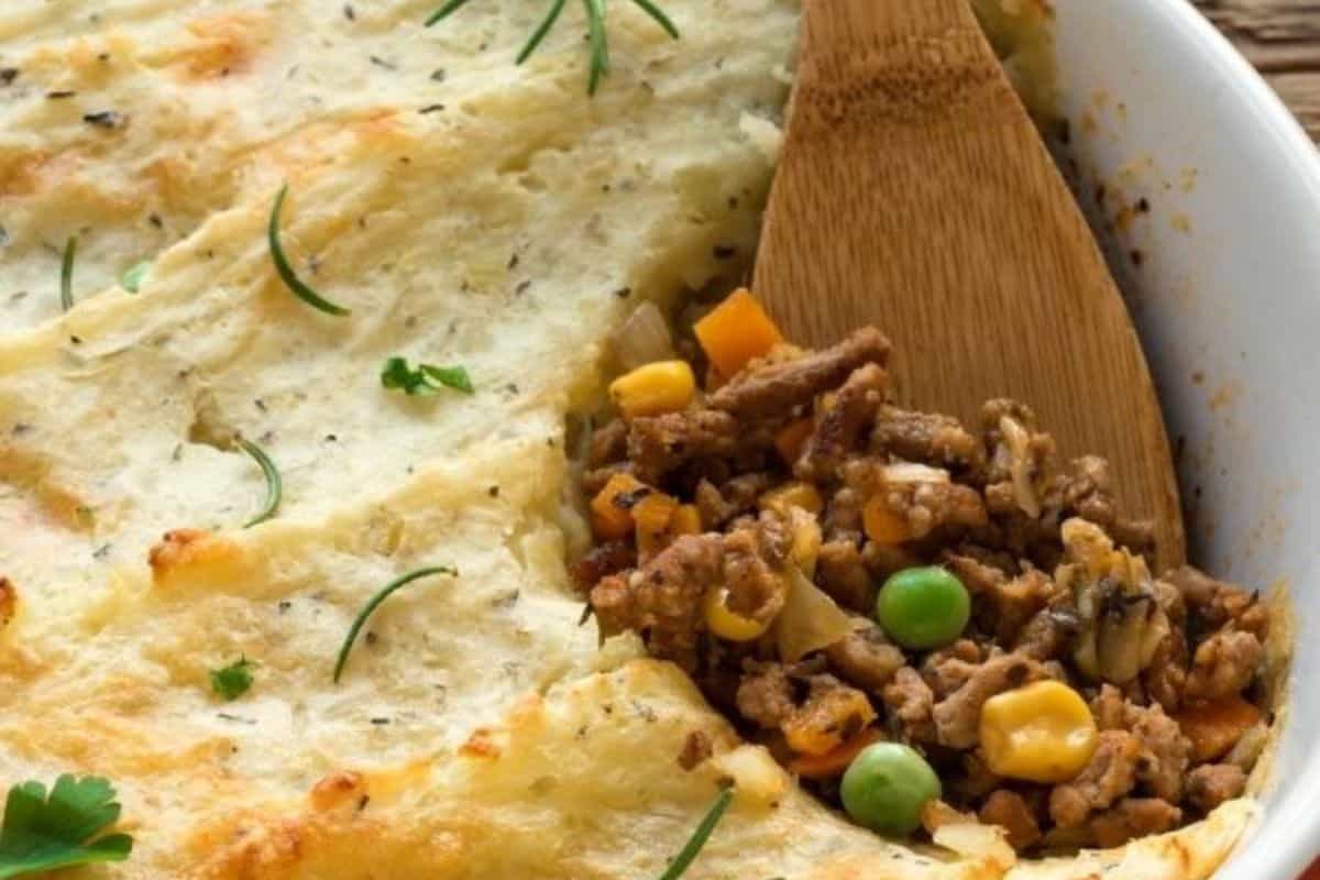 Shepherd's pie with mashed potato topping, revealing a crowd-pleasing ground meat and vegetable filling with peas and corn, garnished with herbs. Perfect for potluck recipes or a St. Patrick's Day celebration. Wooden spoon partially seen.