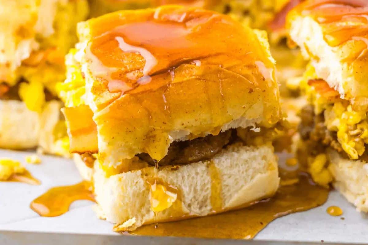 Start your day with a mouthwatering brunch slider featuring crispy bacon, fluffy eggs, and savory sausage on a bun, all topped with a drizzle of maple syrup.
