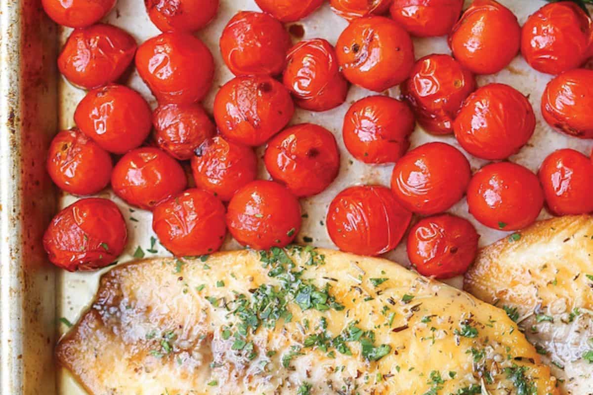 Baked fish fillet topped with herbs and grated cheese beside roasted cherry tomatoes on a baking tray—one of those delicious sheet pan meals that promise easy cleanup.