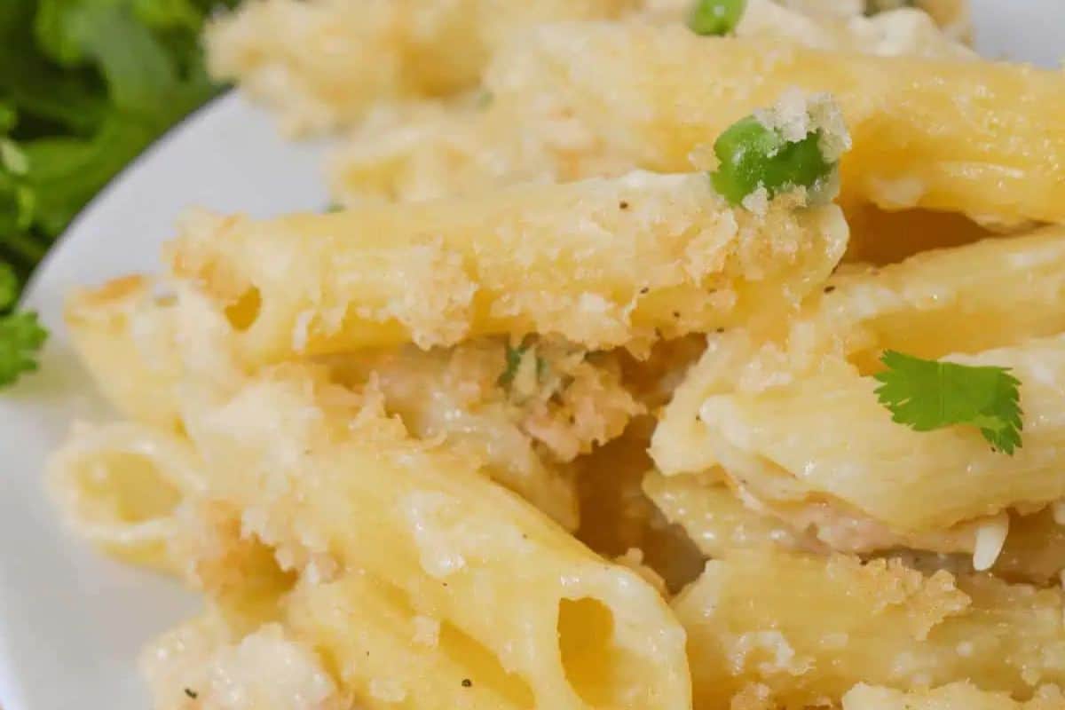 A close-up of a plate of baked garlic parmesan pasta with chicken. The dish features penne pasta coated in a creamy cheese sauce, topped with crispy breadcrumbs and garnished with green peas and fresh parsley.