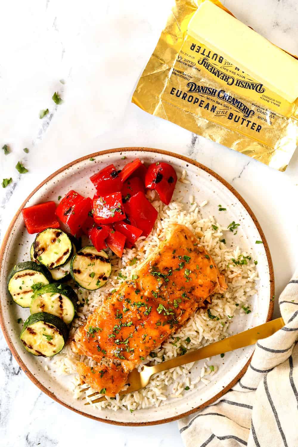 A recipe for a delightful dish: grilled salmon in sauce with white rice, sliced grilled zucchini, and red bell peppers on a plate. Butter wrapper and striped cloth napkin rest on the marble surface, ready to impress at your next potluck.