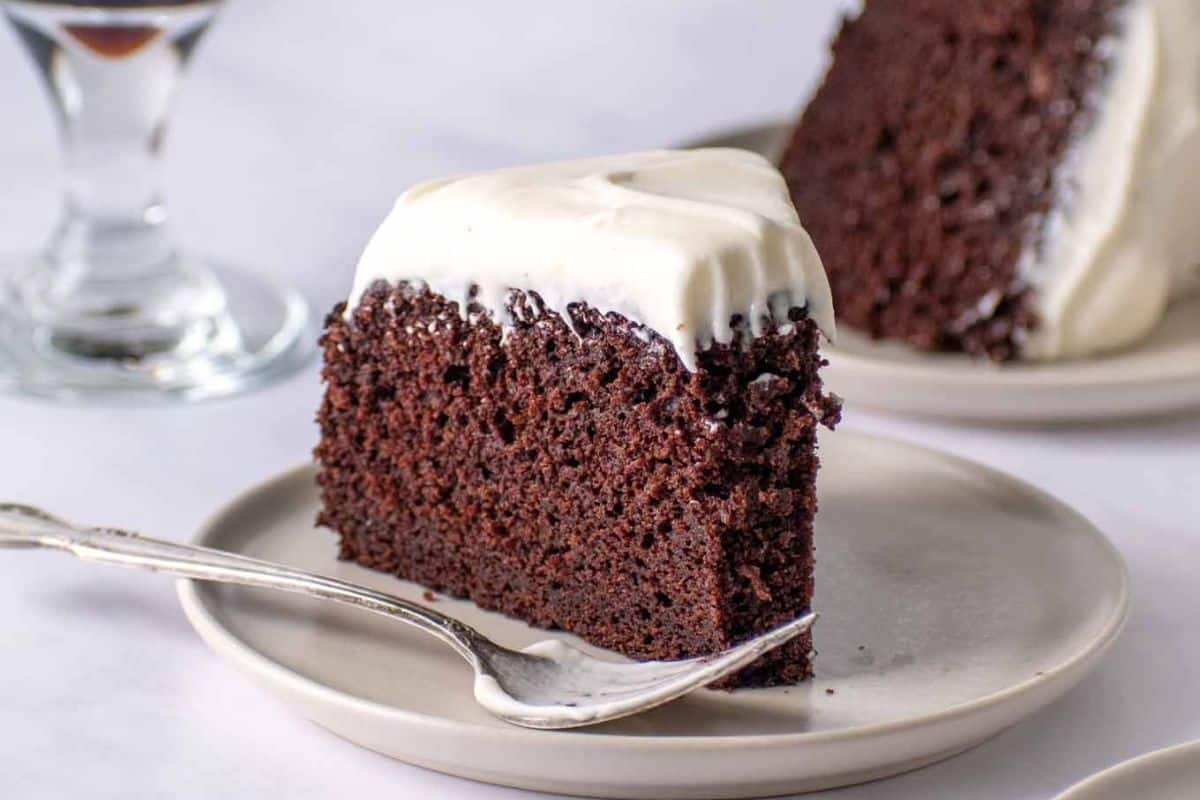 A slice of Guinness chocolate cake with white frosting on a plate evokes the warmth of Irish-inspired comfort food, accompanied by a fork and a glass in the background.