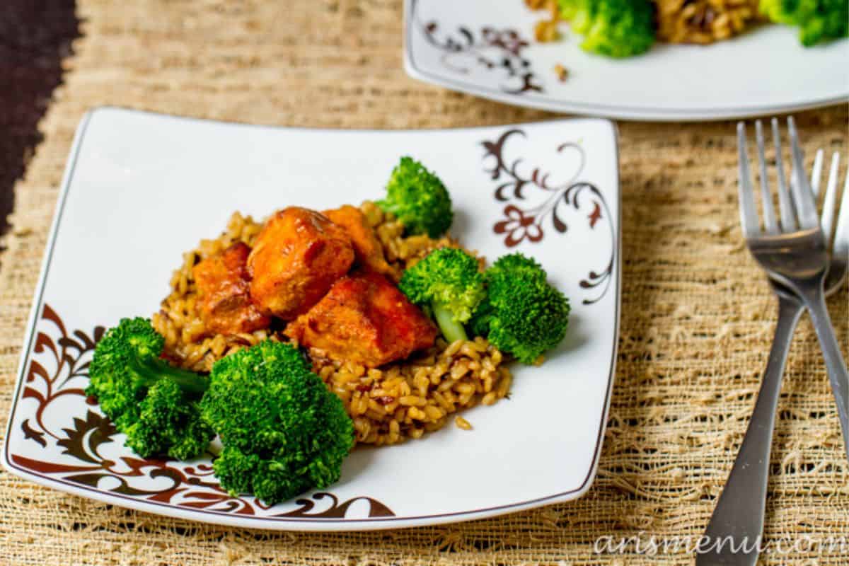 A square plate with rice, broccoli florets, and pieces of glazed chicken sits on a woven placemat, with a fork and knife nearby—perfect for those seeking easy Weight Watchers recipes.
