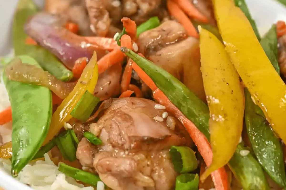 A close-up of delicious stir-fried chicken with snap peas, yellow bell peppers, carrots, and red onions over white rice, topped with sesame seeds—a sheet pan meal for easy cleanup.