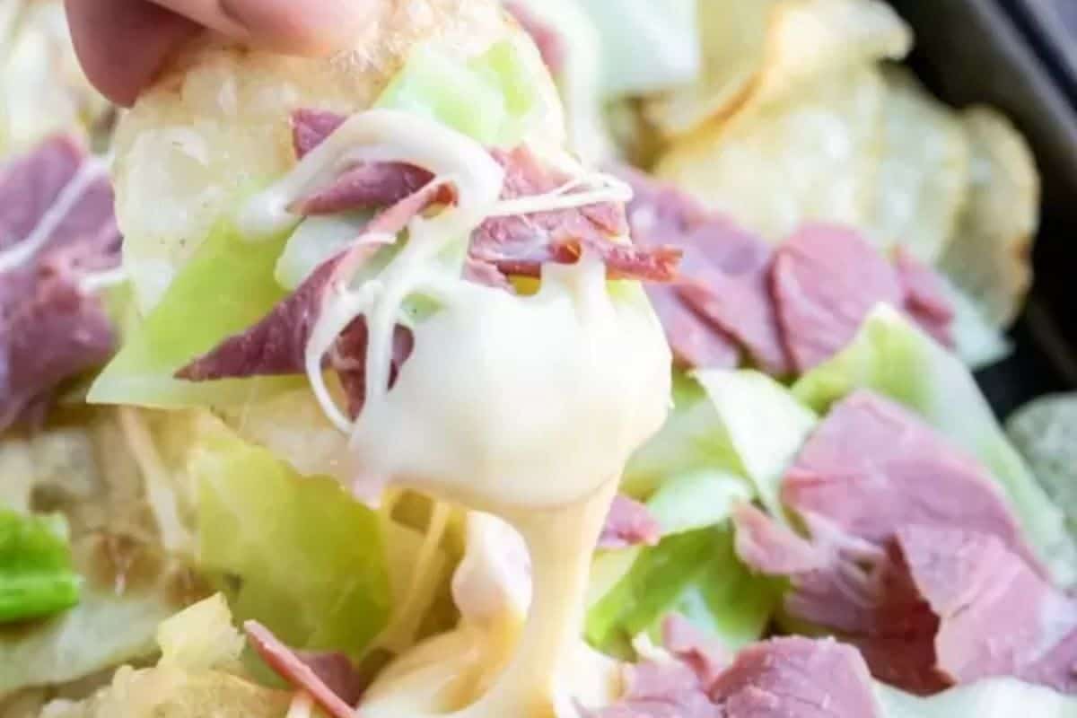 A hand holds a chip draped with melted cheese, shredded corned beef, and cabbage, embodying Irish-inspired nachos. Similar toppings are tempting in the background.