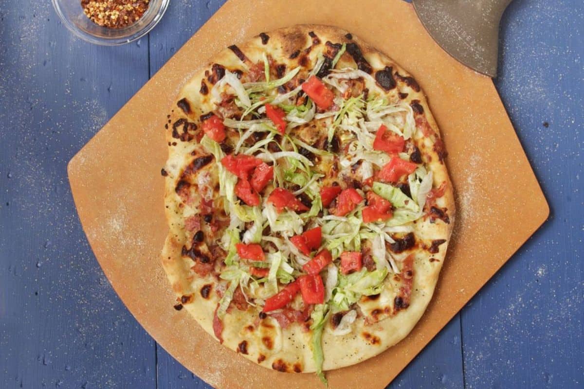 A freshly baked pizza topped with lettuce, tomato, and cheese sits invitingly on a wooden board beside a bowl of chili flakes on a blue surface. It's the perfect night in dish and one of those must-try pizza recipes that promises to delight.