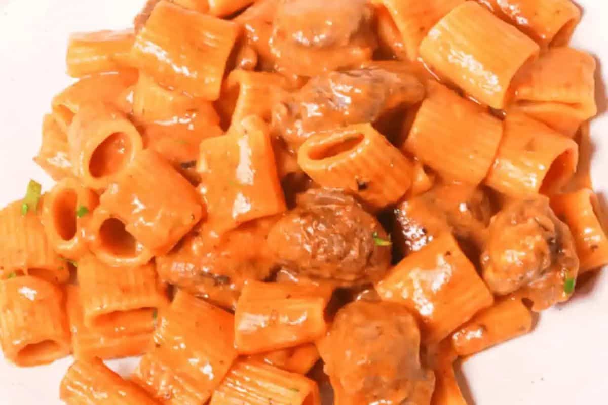 A close-up of Italian pork sausage pasta featuring rigatoni coated in a creamy tomato-based sauce. The dish includes chunks of browned sausage and is garnished with finely chopped herbs.