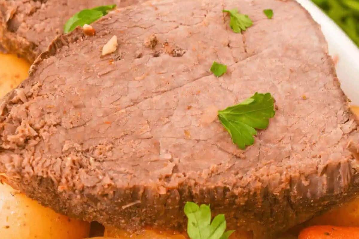 Close-up of a cooked slice of beef pot roast garnished with parsley, served on top of potatoes and other vegetables—a comforting classic perfect to add to your dinner routine or collection of slow cooker recipes.