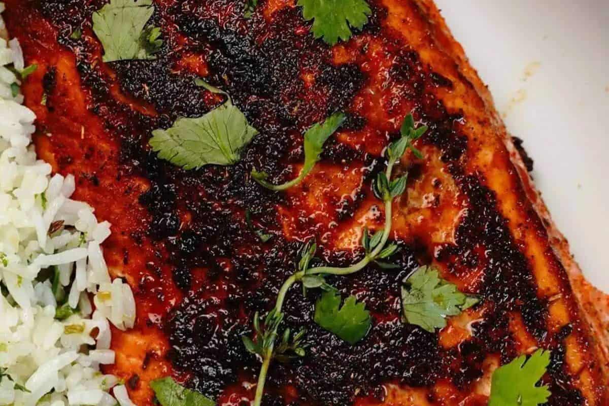 Close-up of an irresistible jerk salmon fillet, perfectly grilled with herbs and topped with a dark glaze, served alongside a portion of white rice garnished with green herbs.