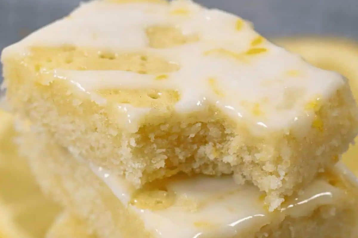 Two lemon-flavored brownies stacked, topped with a luscious lemon glaze. The top bar has a bite taken out, revealing its delightful texture—reminiscent of the finest blondies.