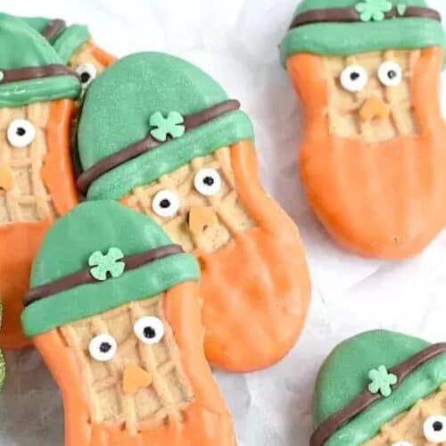 Cute cookies shaped like leprechaun faces, adorned with orange and green icing, small eyes, and clover hats, bring a festive touch to your St. Patrick’s Day sweets spread. They're artfully arranged on a white surface with a glittery green clover decoration.