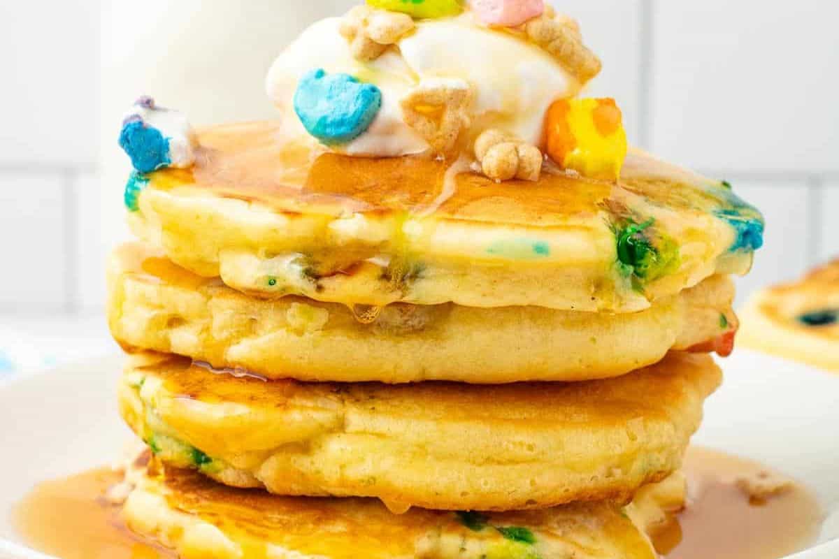 A stack of colorful pancakes with whipped cream and Lucky Charms on top, drizzled with syrup, makes for the perfect brunch delight.
