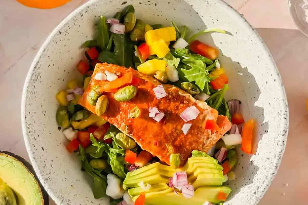 Salmon fillet on a bed of greens with avocado slices, edamame, diced red onions, and bell peppers in a speckled bowl—an irresistible recipe that everyone will love.