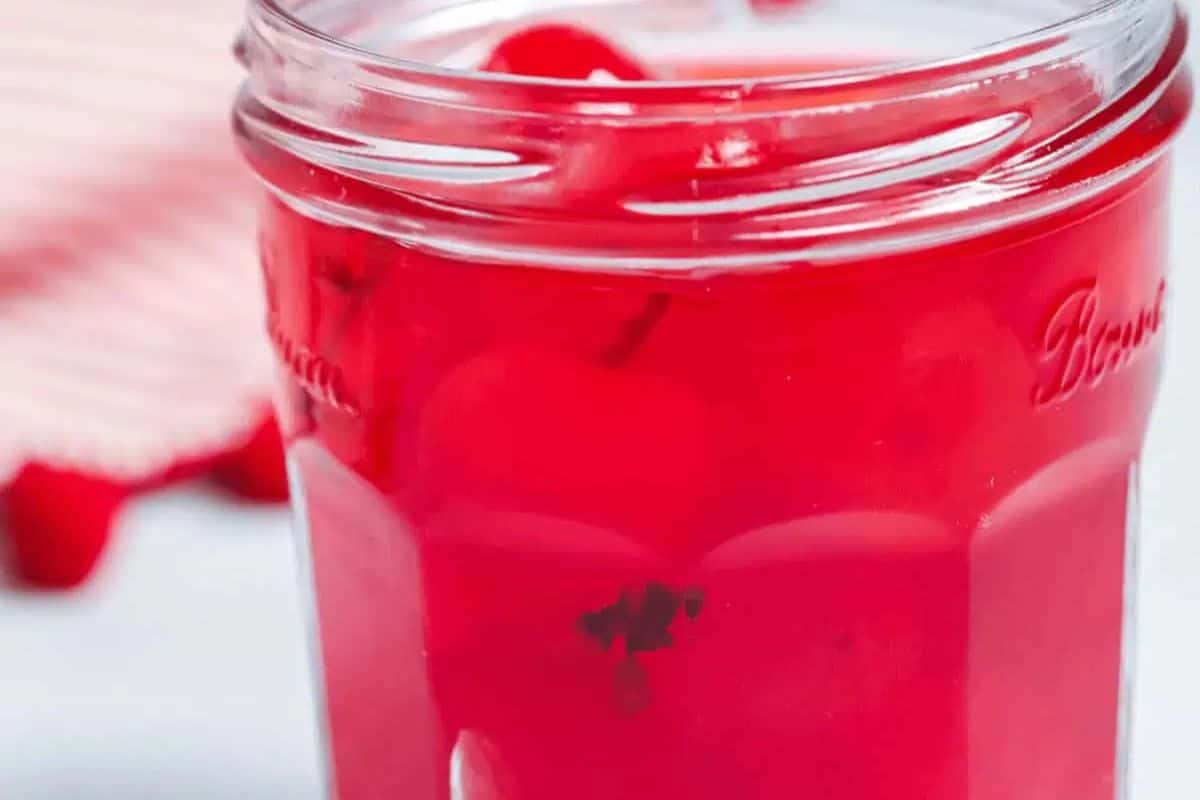 A close-up of a jar filled with moonshine and cherries, perfect for festive recipes, with a striped red and white cloth in the background. Ideal for sharing on Galentine’s Day with your bestie!.