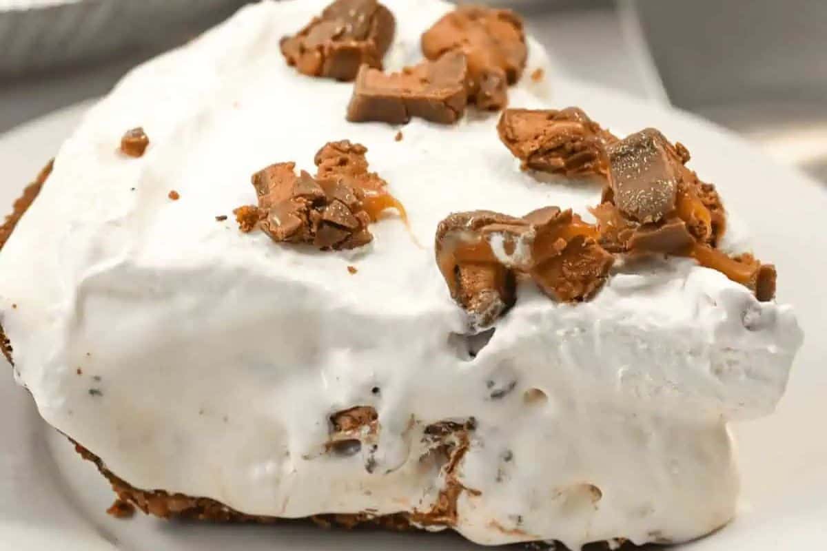 A slice of creamy no-bake carmel pie topped with whipped topping and chopped chocolate pieces rests elegantly on a white plate.