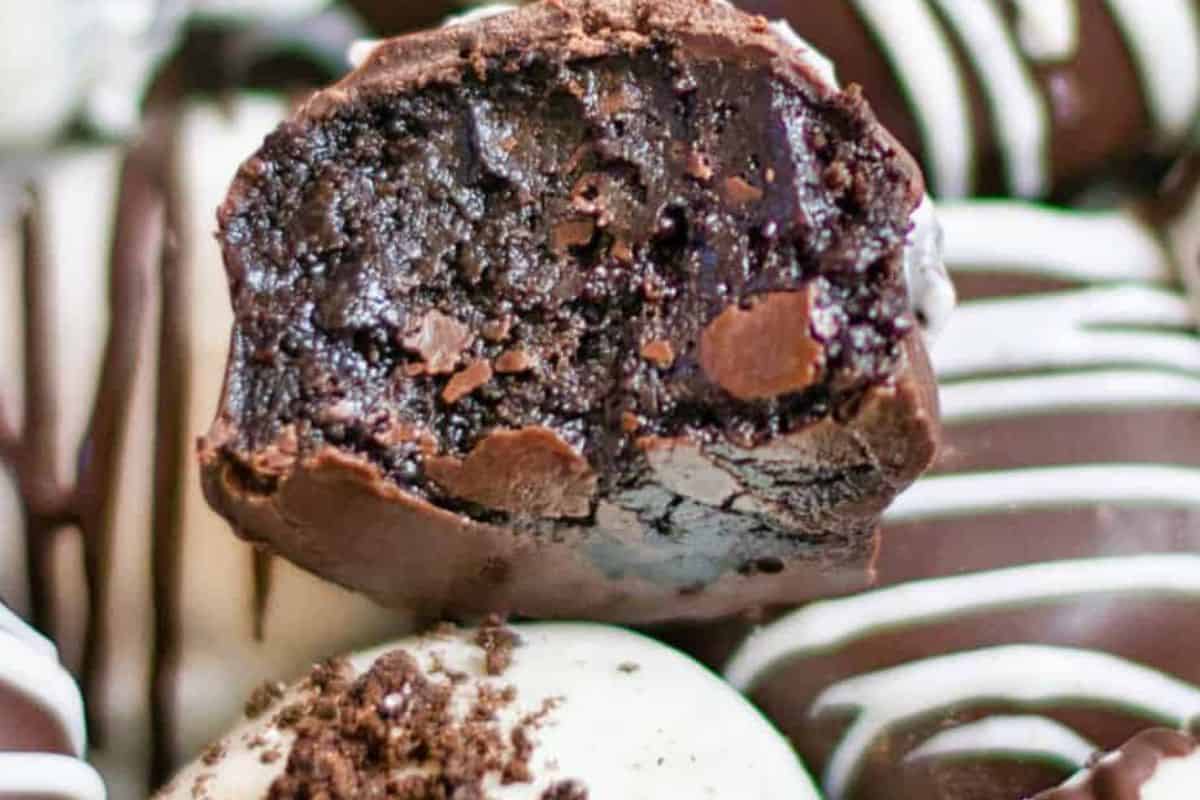 Close-up of a bitten oreo ball with a dense, dark interior. Other truffles, drizzled with white chocolate, are blurred in the background. This no-bake treat is sure to level up your dessert game effortlessly.