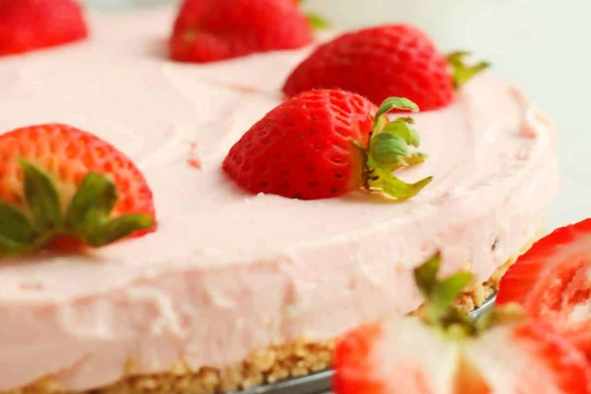 A strawberry-topped, no-bake cheesecake boasts a light pink filling and crumbly crust. Whole strawberries are evenly placed on top, with a sliced strawberry perfectly positioned in the foreground. It's an easy dessert that elevates your dessert game effortlessly.