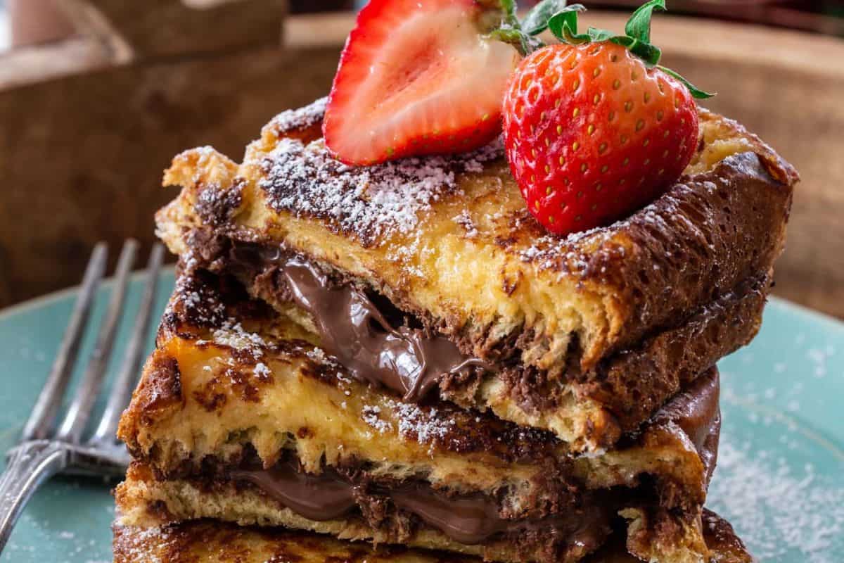 Two slices of French toast filled with chocolate spread, stacked on a plate, topped with halved strawberries and a dusting of powdered sugar—a perfect Valentine’s Day breakfast for those who love sweet and savory brunch recipes.