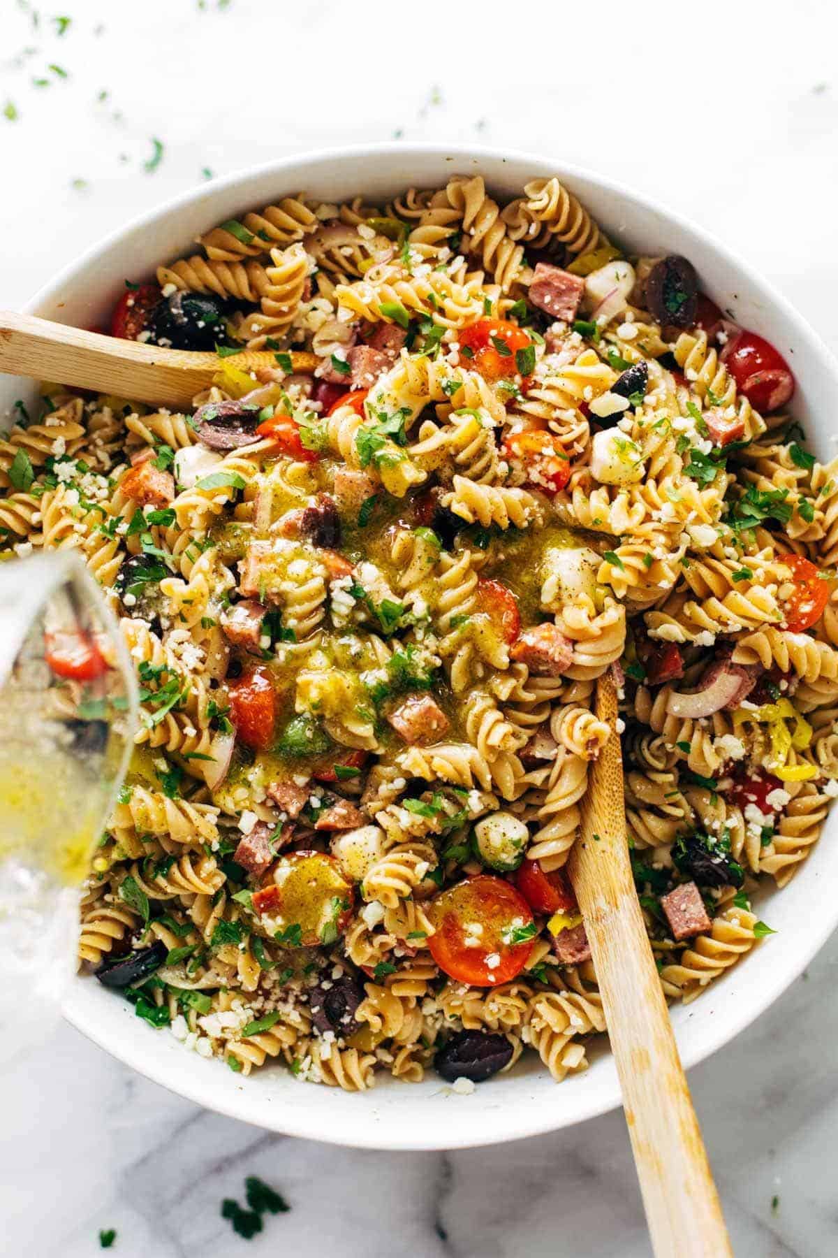 A bowl of pasta salad with rotini, cherry tomatoes, olives, cheese, peppers, and herbs sits ready to impress at any gathering. Two wooden spoons rest in the bowl as a drizzle of dressing is being poured on top—an ideal potluck dish sure to meet recipe requests.