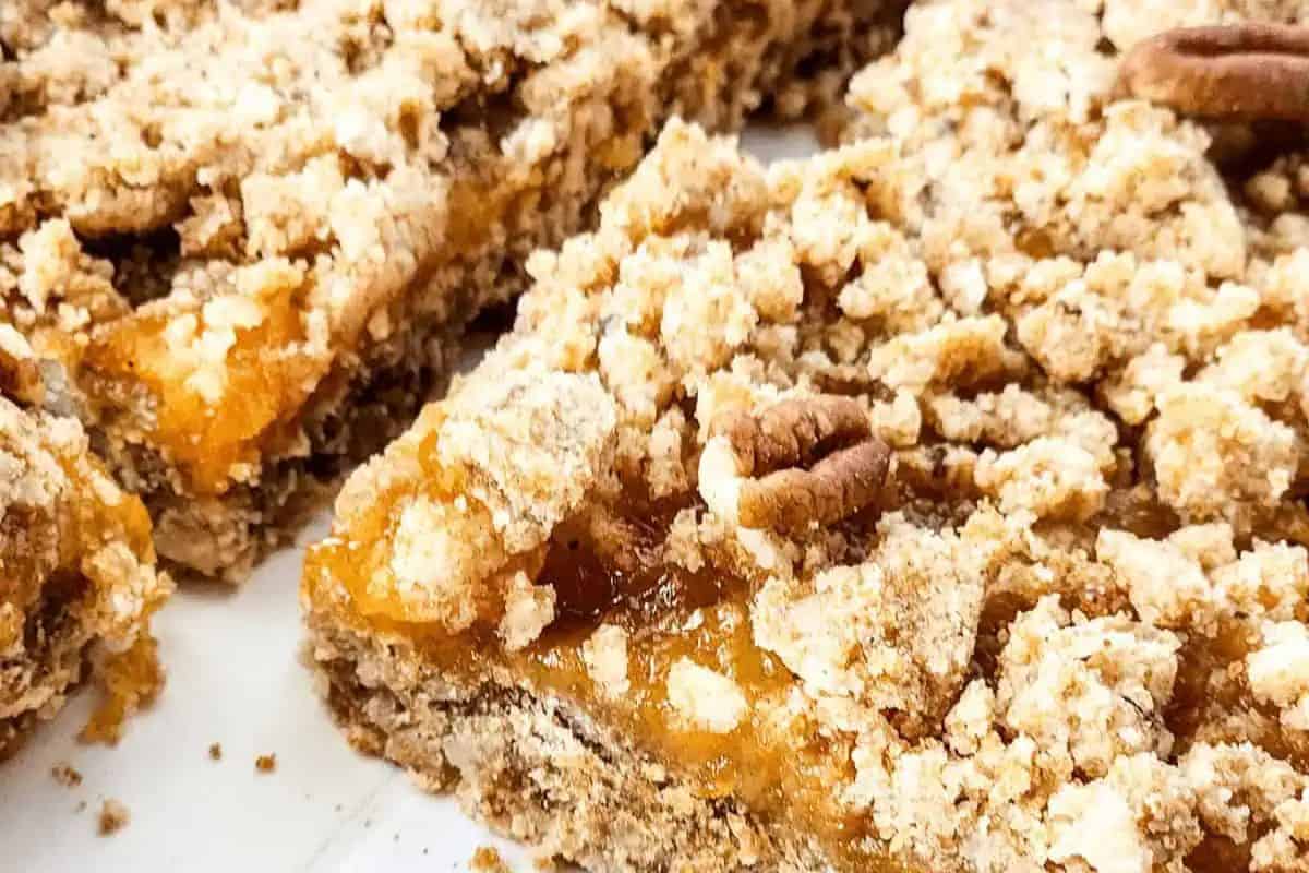Close-up of crumb-topped peach bars on a white surface, featuring a golden apricot filling and a sprinkle of pecans. These delightful treats merge the irresistible texture of blondies with a fruity twist.