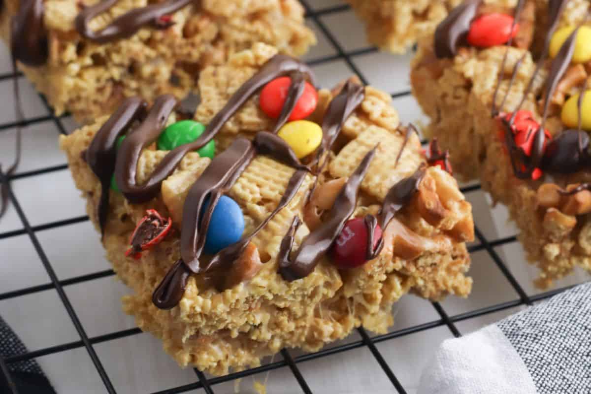 A close-up of a cereal bar, reminiscent of decadent brownies, topped with chocolate drizzle and colorful candy-coated chocolates on a cooling rack.