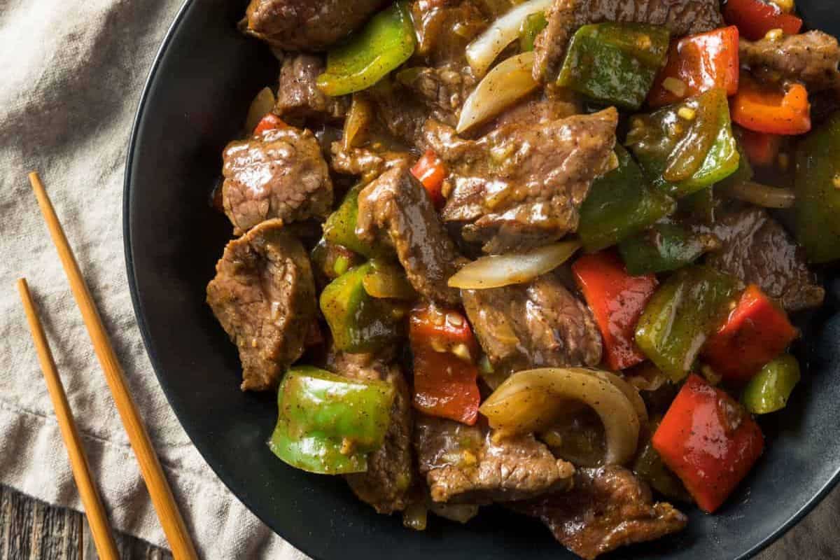 A black plate filled with stir-fried beef, green and red bell peppers, and onions, accompanied by chopsticks on a beige napkin—perfect for easy dinner inspiration or adding to your collection of quick dinner recipes.
