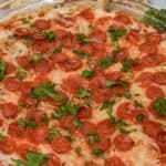 A glass dish filled with Pepperoni Dip, topped with melted cheese and parsley, sits invitingly with slices of bread in the background.