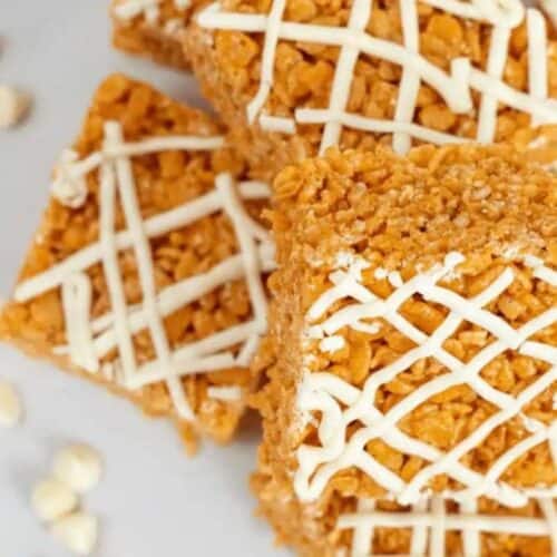 Elevate your snack game with these square peanut butter-flavored cereal bars, elegantly drizzled with white chocolate. Stacked on a pristine white surface, they're accompanied by scattered white chocolate chips, adding a touch of irresistible indulgence.