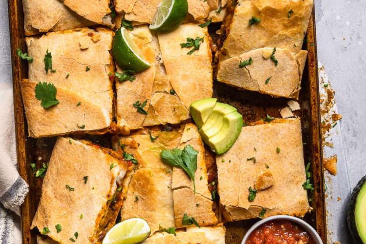 A baking sheet with sliced quesadillas, garnished with lime wedges, avocado slices, cilantro, and a small bowl of salsa on the side—perfect for Sheet Pan Meals that promise delicious flavors and easy cleanup.