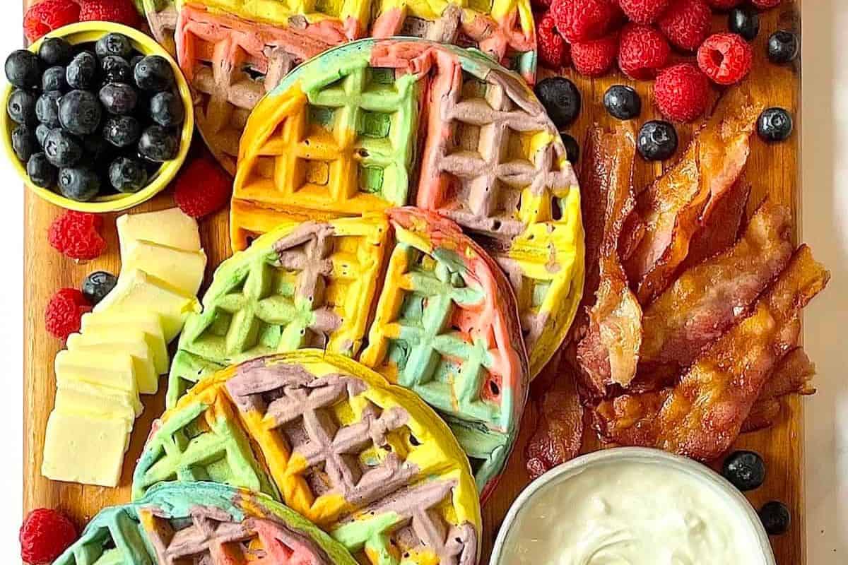 A delightful brunch spread features colorful waffles, crispy bacon, blueberries, raspberries, cheese slices, and creamy dip artfully arranged on a wooden board—perfect for a St. Patrick's Day morning feast.