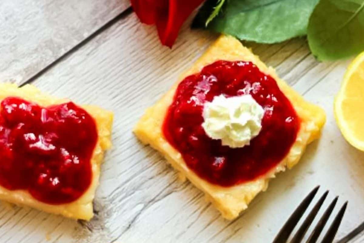 Two square raspberry lemonade bars are topped with red jam and a dollop of cream on a wooden surface. A lemon wedge, fork, and red rose are nearby.
