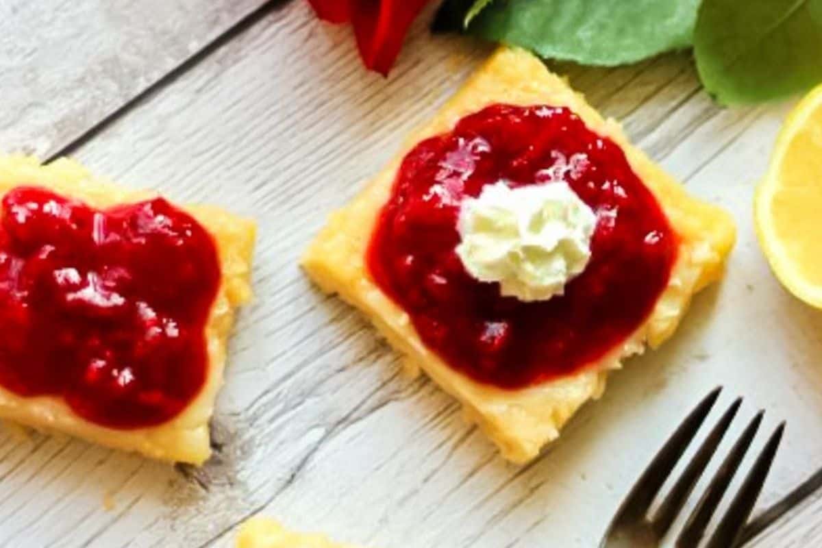 Square raspberry lemondade bars with red fruit topping and a dollop of cream rest on a wooden surface, showcasing the perfect treat for Galentine's Day. Next to them lie a fork and a lemon slice, ready for you and your bestie to indulge.