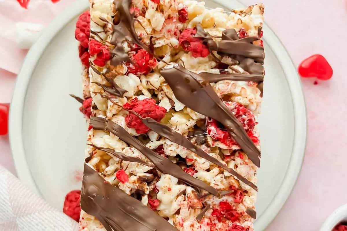 A rice krispie treat with raspberries and rice crisps, drizzled with chocolate on a white plate adorned with scattered heart-shaped decorations. Elevate your snack game by exploring various Rice Krispie Treat recipes to complement this delightful creation.