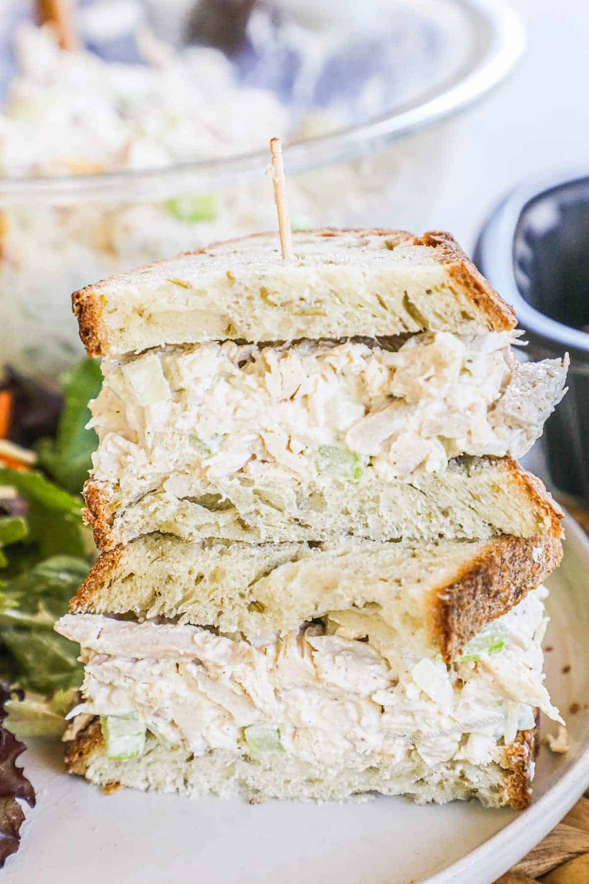 Foolproof chicken salad sandwich stacked on whole grain bread with a toothpick. Perfect for potluck dishes, it's served on a plate with mixed greens on the side.