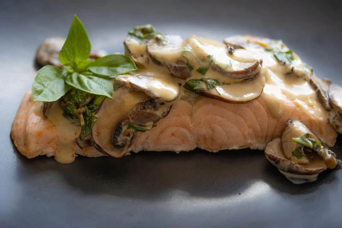 Irresistible salmon fillet topped with creamy mushroom sauce, garnished with fresh basil leaves, served on a dark plate.