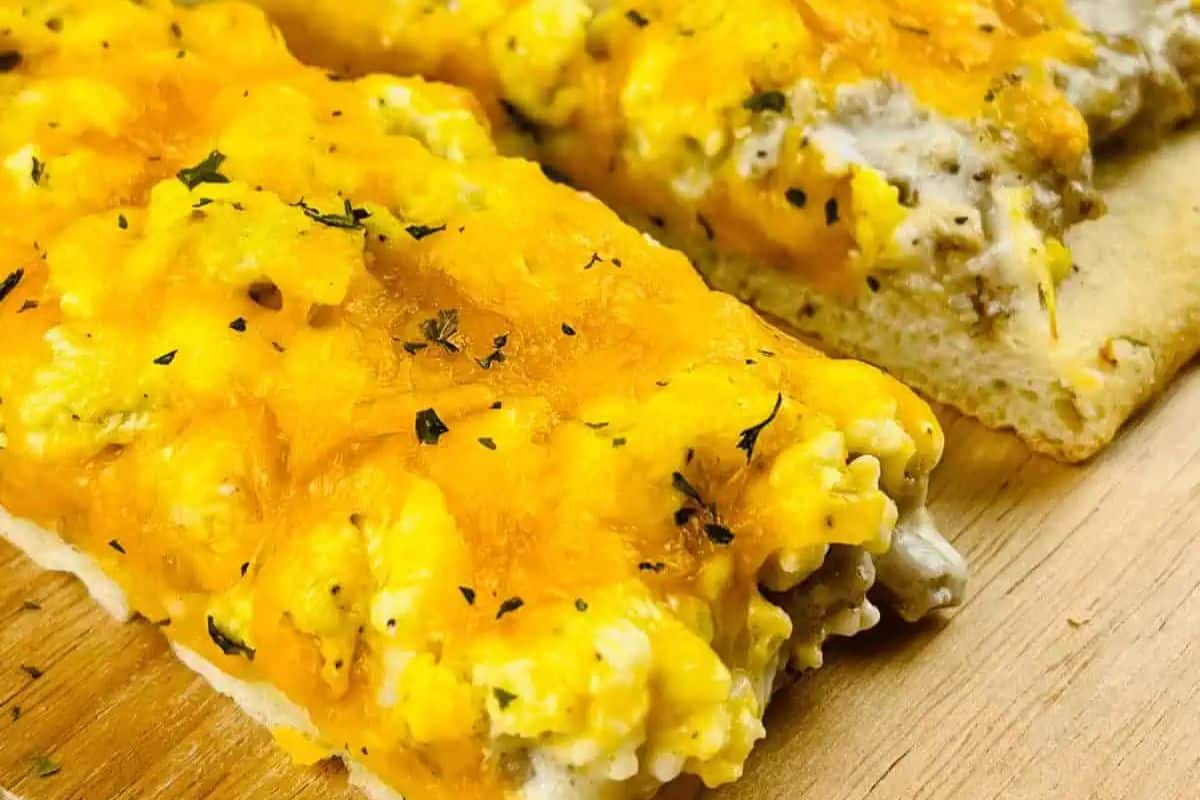 Close-up of two slices school breakfast pizza topped with scrambled eggs, melted cheese, herbs, and sausage pieces on a wooden surface—perfect for a cozy night in.