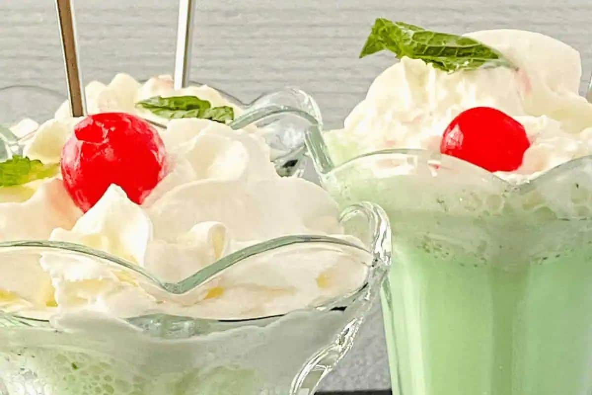 Two cute glass cups of mint milkshake, perfect for St. Patrick’s Day, topped with whipped cream, cherries, and mint leaves.