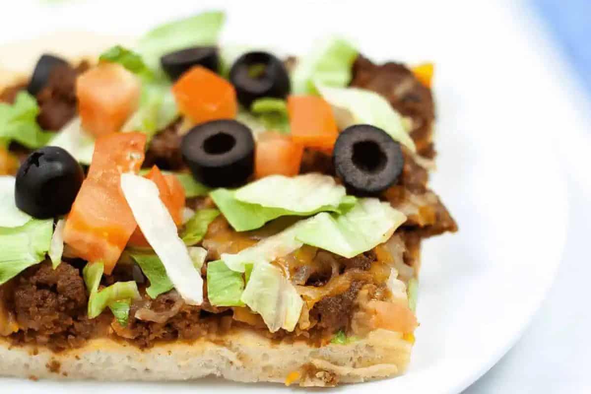 A slice of taco pizza, a must-try delight, features ground beef, cheese, lettuce, tomato, and black olives on a white plate—perfect for a cozy night in with the best pizza recipes.
