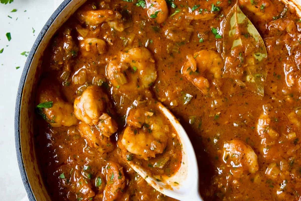 A pot of restaurant-quality shrimp creole brimming with shrimp, sauce, and seasonings, all garnished with fresh herbs. A white spoon sits invitingly in the dish.
