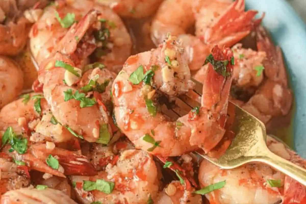 A fork holds a succulent, restaurant-quality shrimp scampi garnished with chopped parsley and spices. Other delectable shrimp dishes are visible in the background on a plate, promising an enticing culinary experience.
