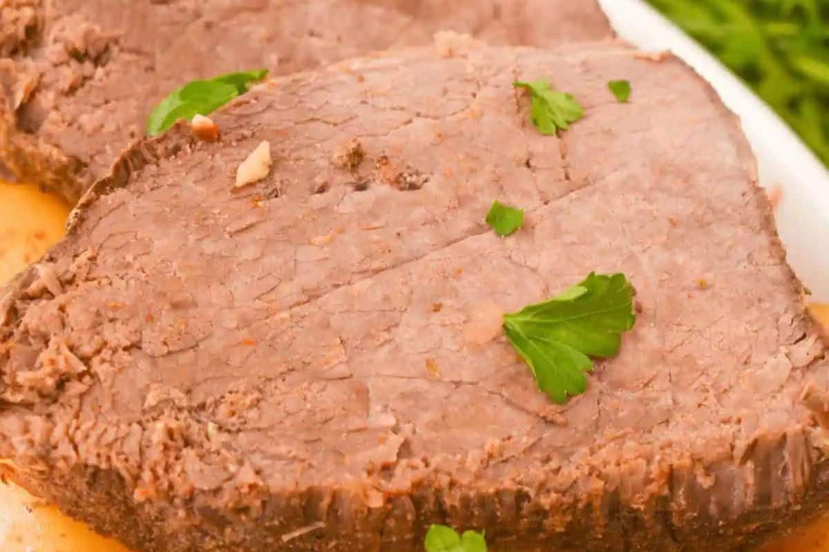 A close-up of two slices of slow-cooked beef sirloin tip roast on a white plate. The meat appears tender and juicy, with a slightly browned exterior and garnished with fresh parsley.