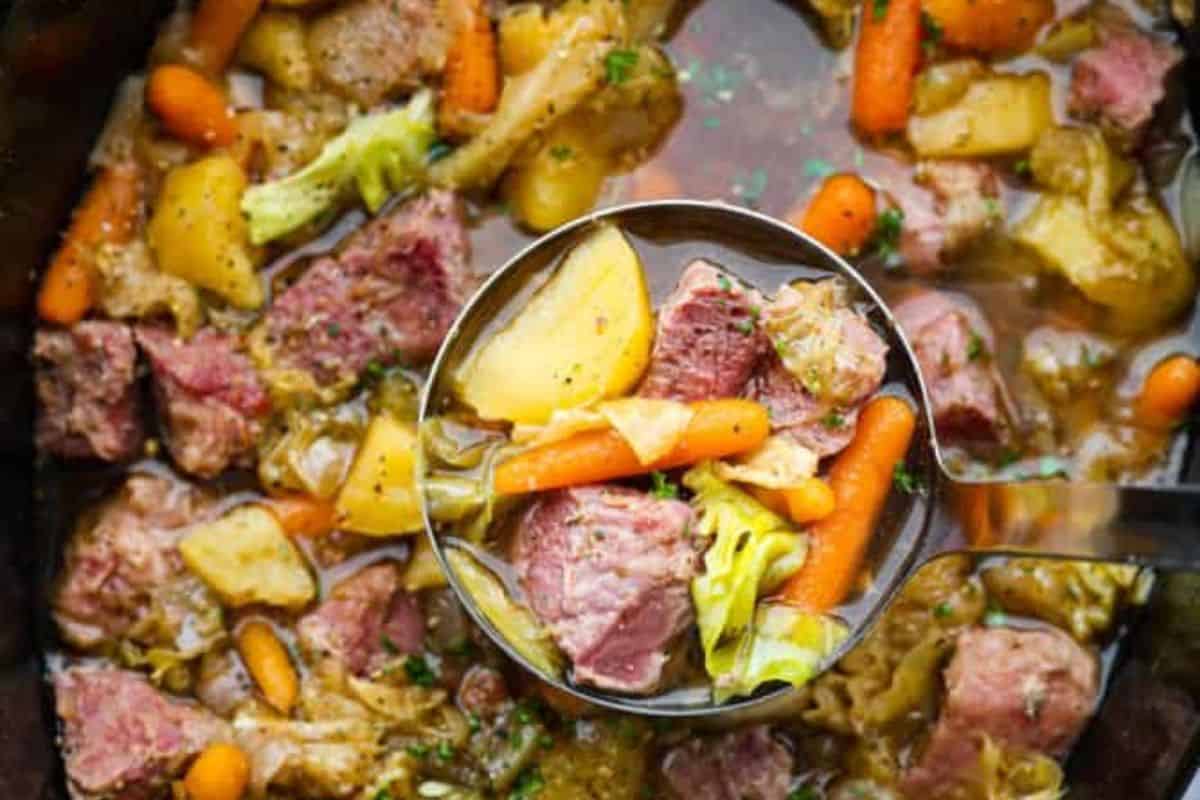 A ladle holds a serving of stew with corned beef, potatoes, carrots, and cabbage, reminiscent of traditional recipes. The hearty mix is surrounded by more stew in a pot, bringing to mind the comforting flavors of classic corned beef and cabbage dishes.