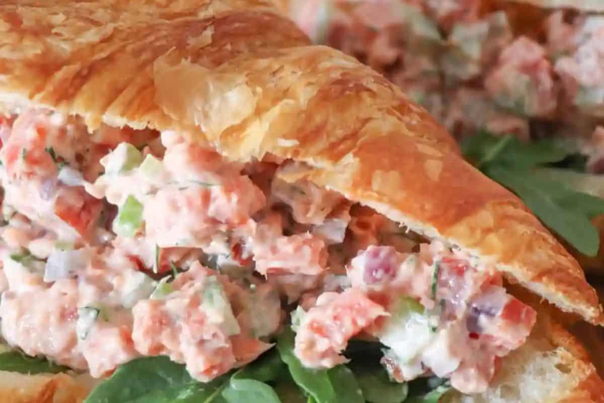 Irresistible close-up of a flaky croissant filled with a creamy salad mix, including diced vegetables and salmon, set atop a bed of fresh greens.