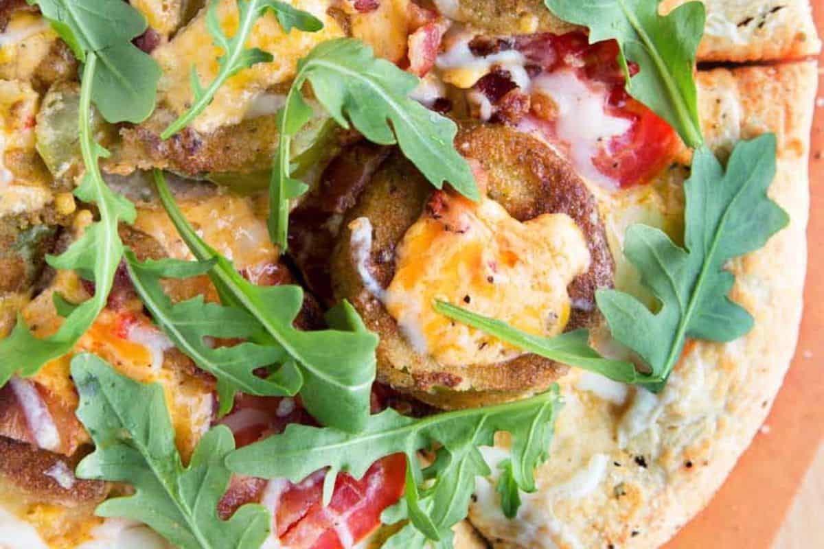 Close-up of a delightful pizza recipe featuring fried green tomatoes, melted cheese, sliced tomatoes, and fresh arugula on a wooden surface–ideal for the perfect night in.