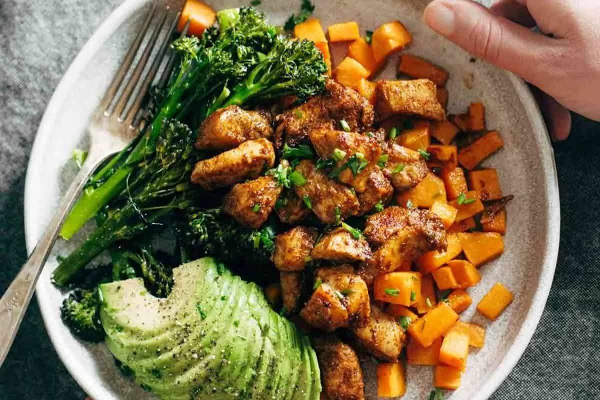 A plate with seasoned chicken pieces, roasted sweet potatoes, broccolini, and sliced avocado—an inviting meal that's perfect for those seeking easy Weight Watchers recipes. A fork and a hand are visible holding the plate.