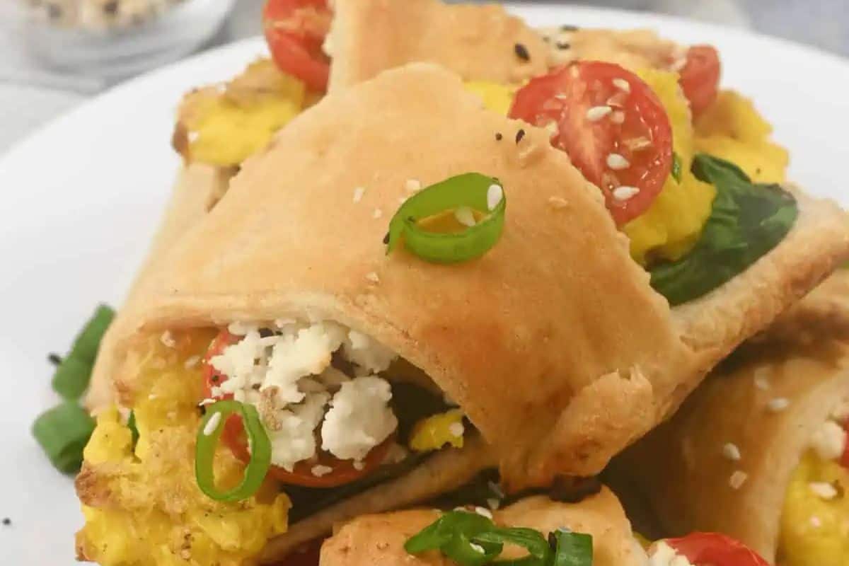 Golden crescent roll stuffed with scrambled eggs, spinach, tomatoes, and crumbled cheese, garnished with green onions and sesame seeds—a sweet and savory delight perfect for brunch recipes.
