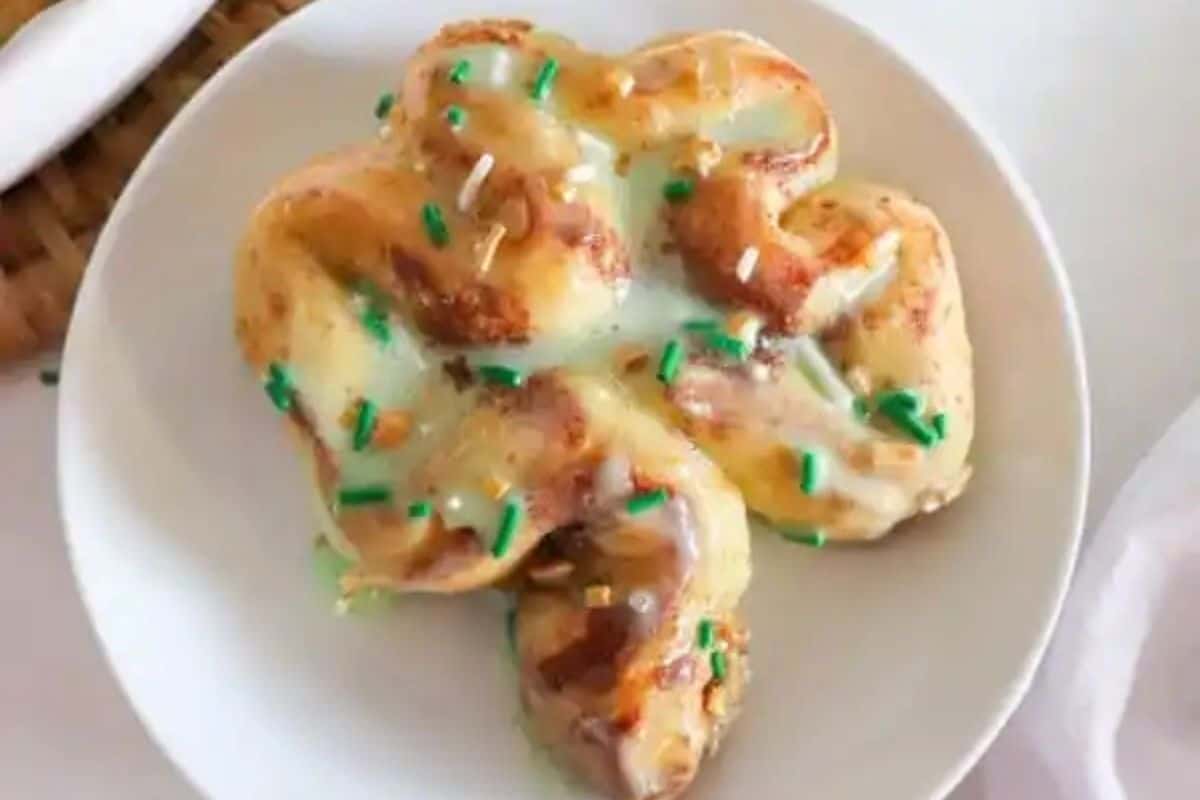 A cinnamon roll shaped like a clover, topped with white icing and green sprinkles, sits invitingly on a white plate, perfect for a festive St. Patrick's Day brunch.