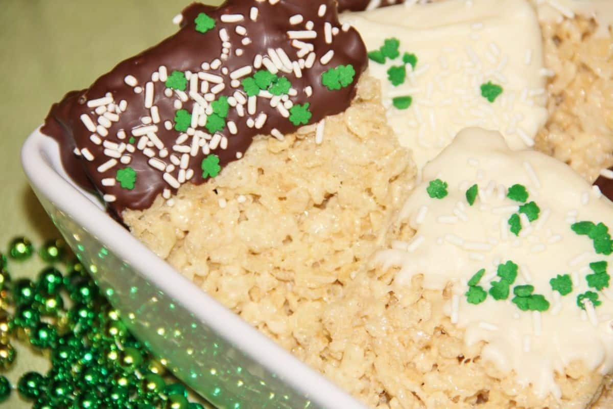 Rice Krispie treats partially dipped in chocolate, decorated with cute green and white sprinkles, sit in a dish. Perfect for St. Patrick's Day celebration, green bead decorations are nearby on a light green surface.