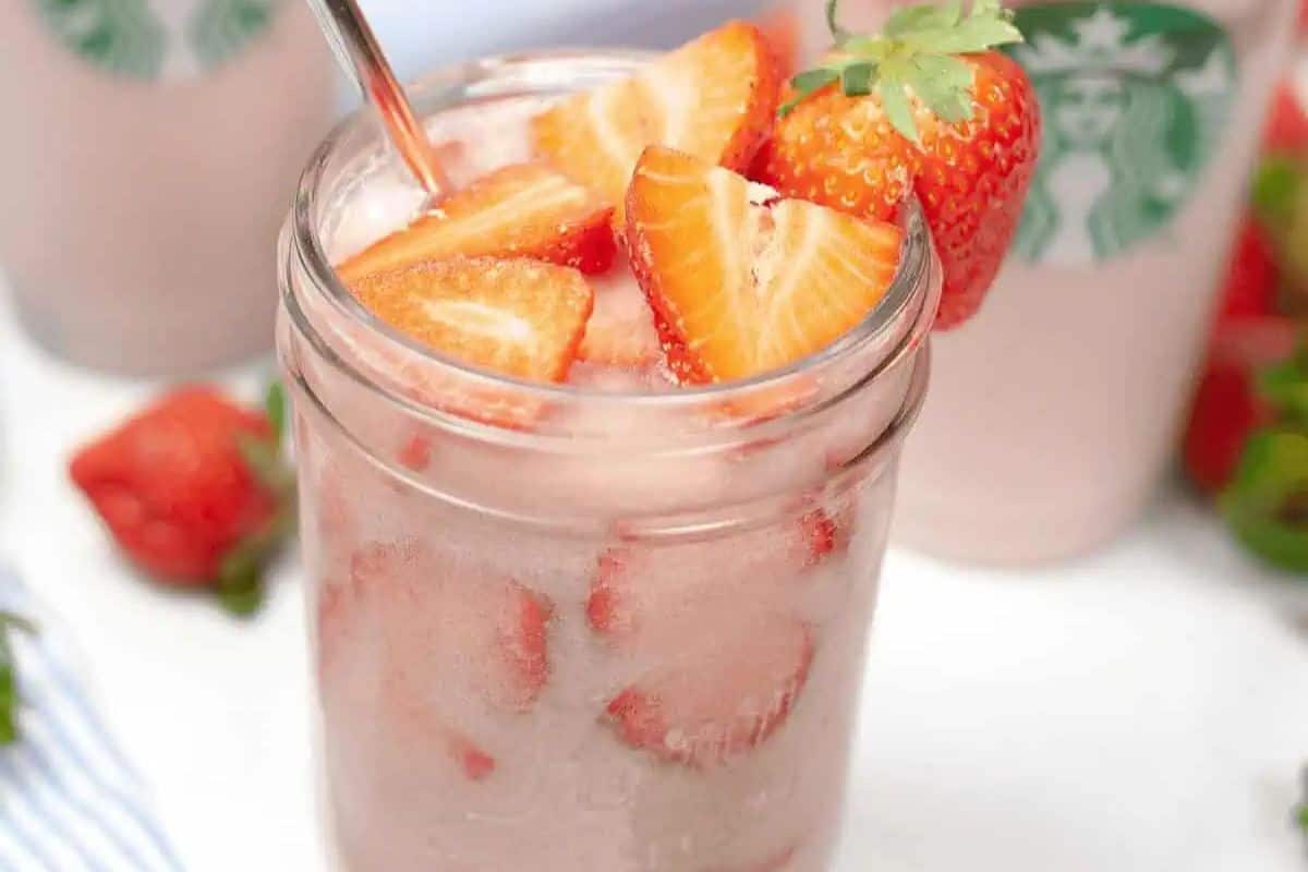 A glass of iced pink drink, perfect for Galentine’s Day, is topped with sliced strawberries and garnished with a whole strawberry on the rim. Starbucks cups are visible in the background, setting the scene for a festive celebration with your bestie.